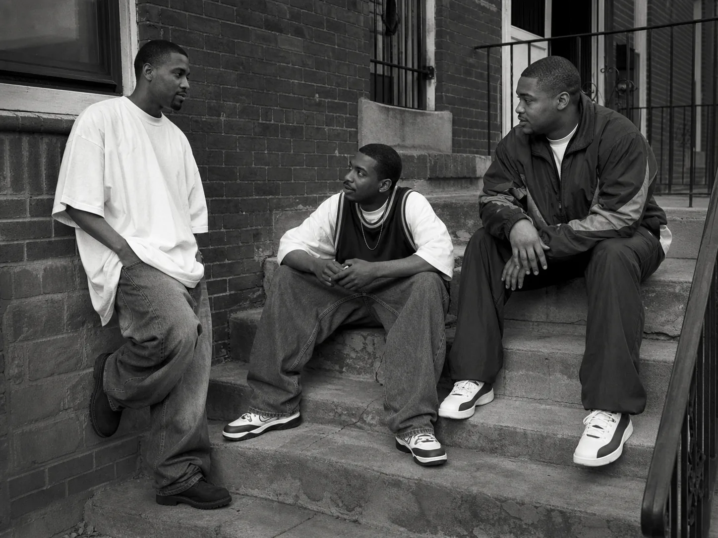 Three Black men wearing baggy denim, a layered sports jersey and a nylon tracksuit on apartment steps