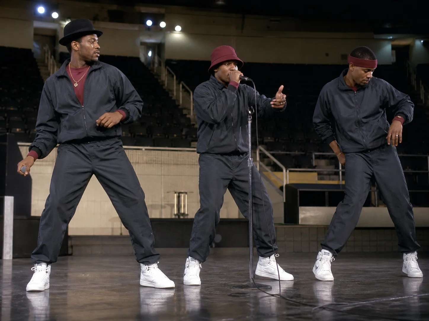 Coordinated late-1980s rap group stage outfits Fictional Black rap trio rehearsing in coordinated charcoal athletic outfits with muted burgundy accents and white high-top sneakers