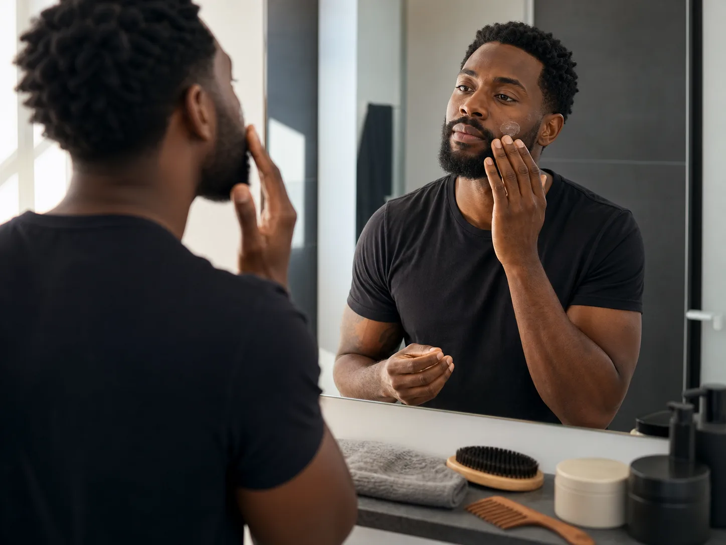 Simple non-medical Black men's grooming routine Black man with natural skin texture applying plain moisturizer beside beard brush, comb, towel and unlabeled containers at bathroom mirror